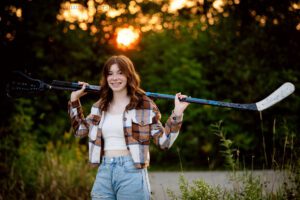 senior pictures in milwaukee. high school senior girl wearing jeans and brown flannel shirt. standing in park hold a lacrosse stick and a hockey stick.