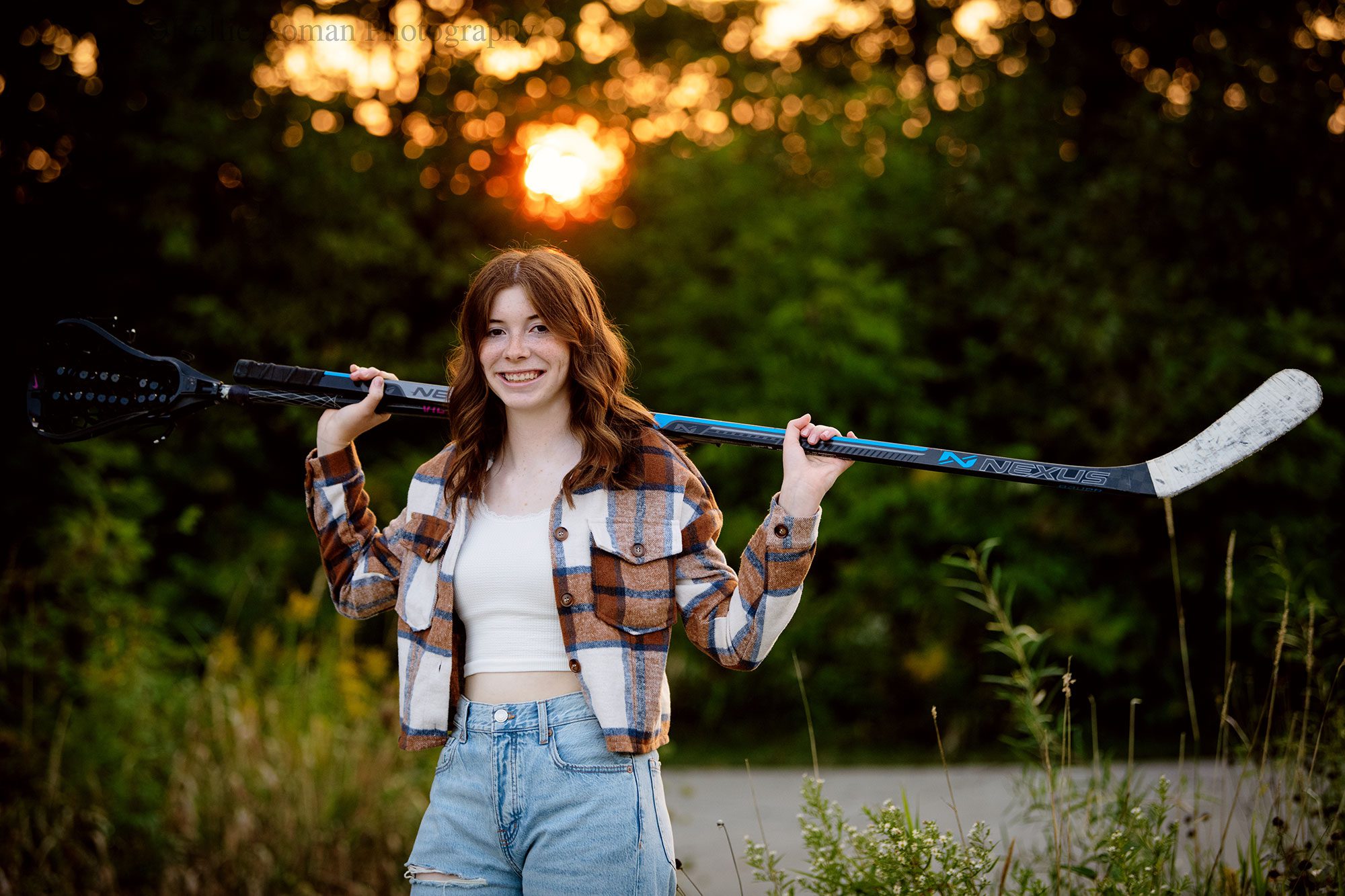 senior pictures in milwaukee. high school senior girl from Muskego is wearing jeans and brown flannel shirt. she has a hockey and lacrosse sticks over her shoulders while she's smiling at the camera. 
