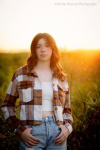 senior pictures in milwaukee. senior girl standing with thumbs in pocket of jeans. she has a brown flannel sweater on and brown hair. the sun is setting gold on the horizon behind her. she's looking at the camera with a serious face.
