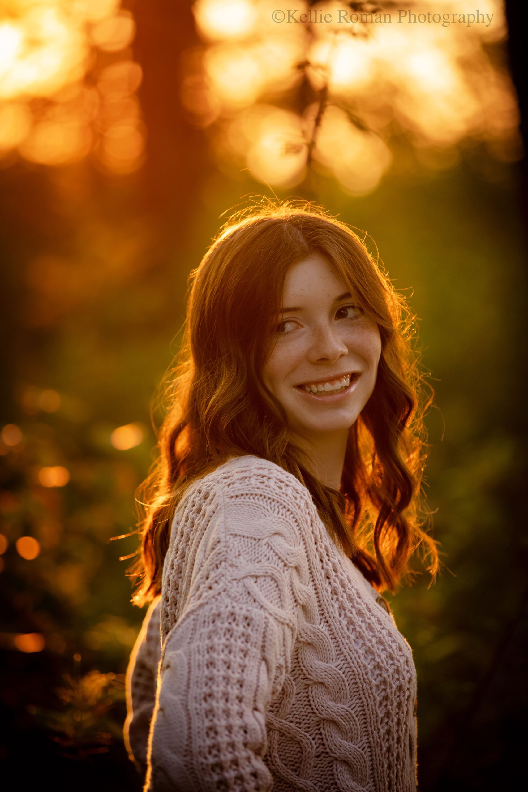 senior pictures in milwaukee. high school Muskego senior is in milwaukee parking with trees and golden setting sun behind her. she's looking over her shoulder with her hands on her hips and the sunlight hitting her brown hair. she has a pink knit sweater on. 
