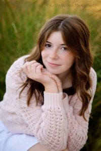 senior pictures in milwaukee. high school senior girl with a pink knit sweater is leaning forward on her knee with her hands under her chin. she is slightly smiling and has brown hair. the background is green.