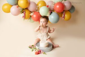 floral cake smash milwaukee. one year old girl sitting on cream backdrop with white cake infront of her. cake is resting on a metal stand, with flowers next to it. girl is wearing cream romper and is putting cake in her mouth. backdrop is balloon garland with purple, yellow, green, blue and red balloons.