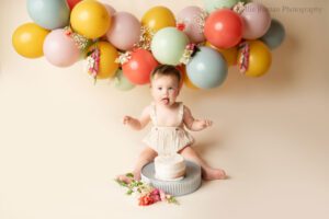floral cake smash milwaukee. one year old girl sticking her tongue out sitting on floor behind white cake on a metal stand. flowers are on the floor by the cake stand. backdrop is cream, with a balloon garland in pink, purple, blue, green and yellow.