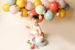floral cake smash milwaukee. one year old touching a white frosted cake resting on a metal stand. girl has a cream romper on. backdrop is cream with a balloon garland or pink, purple, blue, green and flowers.