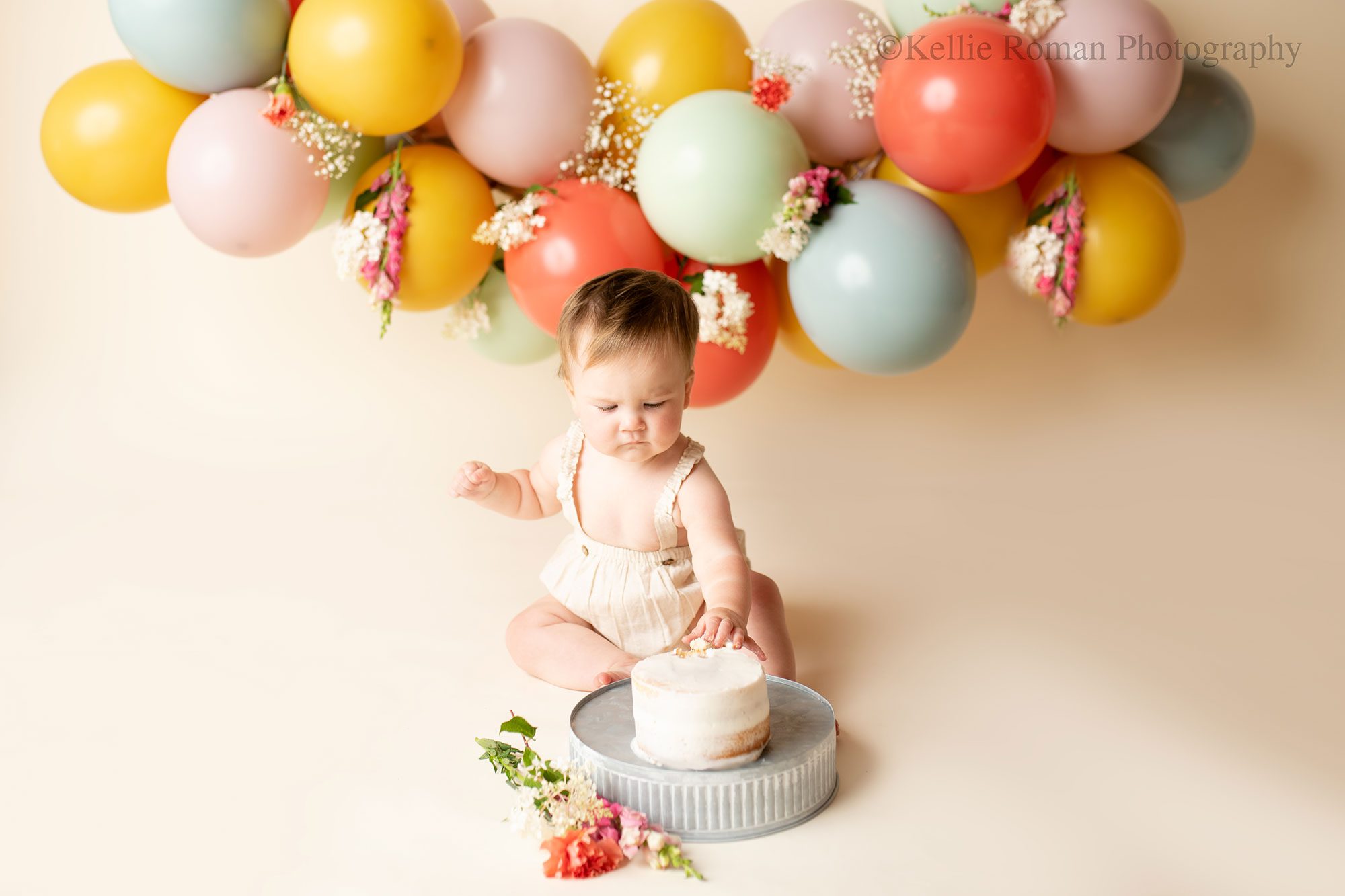 floral cake smash milwaukee. one year old girl sitting on the ground touching a white frosted cake that is on a metal cake stand. there is a cream backdrop with balloon garland behind her. the balloons are blue, green, red, yellow, and purple with fresh flowers on them. 
