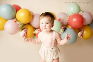floral cake smash milwaukee. one year old girl standing and slightly smiling,. she has blue eyes, and cream romper on. the backdrop is green with a balloon garland that is blue, yellow, purple, red and green. it has flowers on the balloon garland.