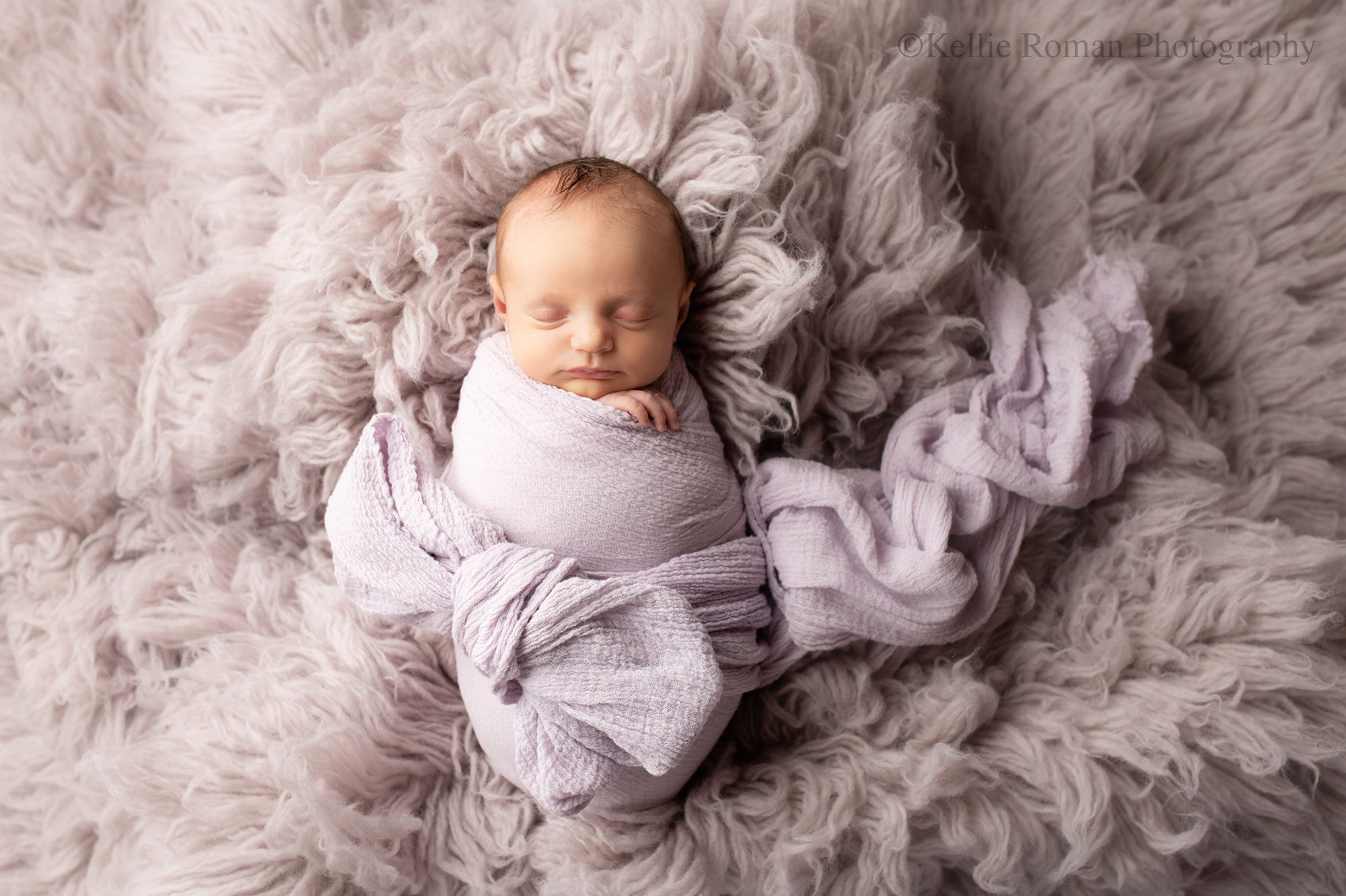 adorable newborn sister. newborn baby in a lavender swaddle that is tied in a bow is sleeping on her back on top of a lavender fluffy rug. she has one hand sticking out of the wrap.