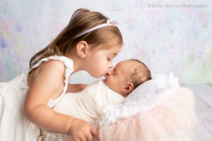 adorable newborn sister. newborn girl is sleeping on her back and swaddled in white wrap. she's in a basket filled with white and pink stuffing. her toddler sister is holding onto the basket and leaning in to give her newborn sister a kiss on the nose.