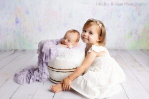 adorable newborn sister. newborn baby is in a white wood bucket sleeping with her hands under her chin. the bucket has ruffle layers of purple and white. her big sister is sitting on the floor with her legs around the bucket and is looking and smiling at the camera. big sister has a white dress on. the backdrop is pastel shades or purple pink and teal.