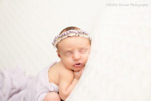 adorable newborn sister. newborn girl sleeping on her belly with her hand under the side of her cheek. she's onto of cream colored fabric with light purple fabric over her diaper. she has a light purple bead headband on.