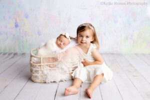 adorable newborn sister. newborn girl swaddled in a white wrap is sleeping on her side in wood basket stuffed with purple and white fabrics. her big sister is sitting next to the basket on the floor with her arm around the basket. the older sister is barefoot and wearing a white dress.