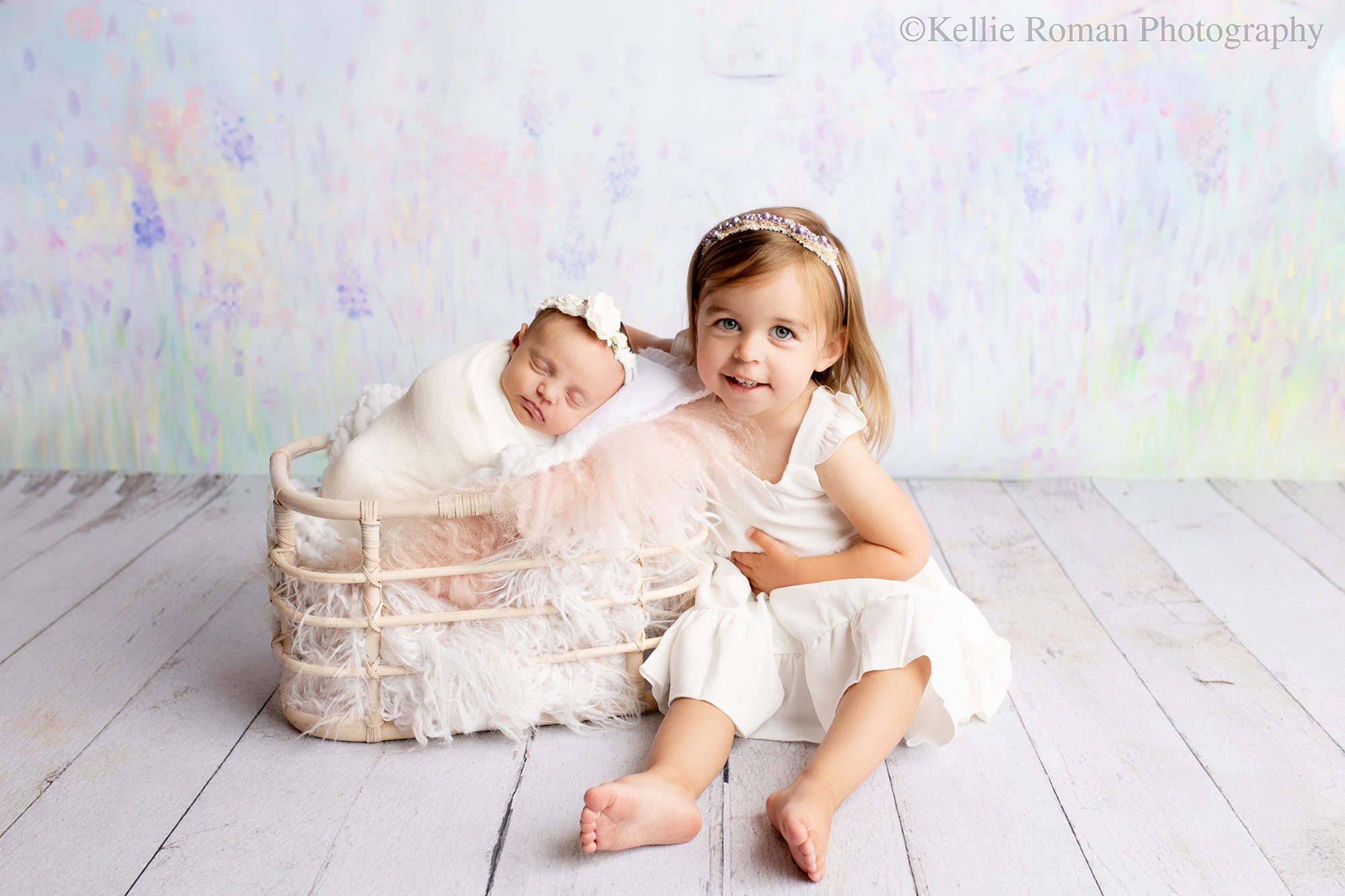 adorable newborn sister. a newborn baby girl is wrapped in a cream fabric and sleeping in a basket with pink and white furs. toddler big sister who is wearing white dress is sitting next to the basket. backdrop is shade for purple 