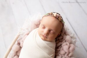 adorable newborn sister. newborn girl swaddled In a white wrap with purple floral headband. she's asleep in a wood basket with white and purple furs.