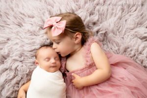 adorable newborn sister. milwaukee newborn is swaddled in white wrap laying on a purple fuzzy rug. toddler sister is laying next to her with a pink fluffy dress on and her arm under her baby sister. big sister is kissing baby on the head.