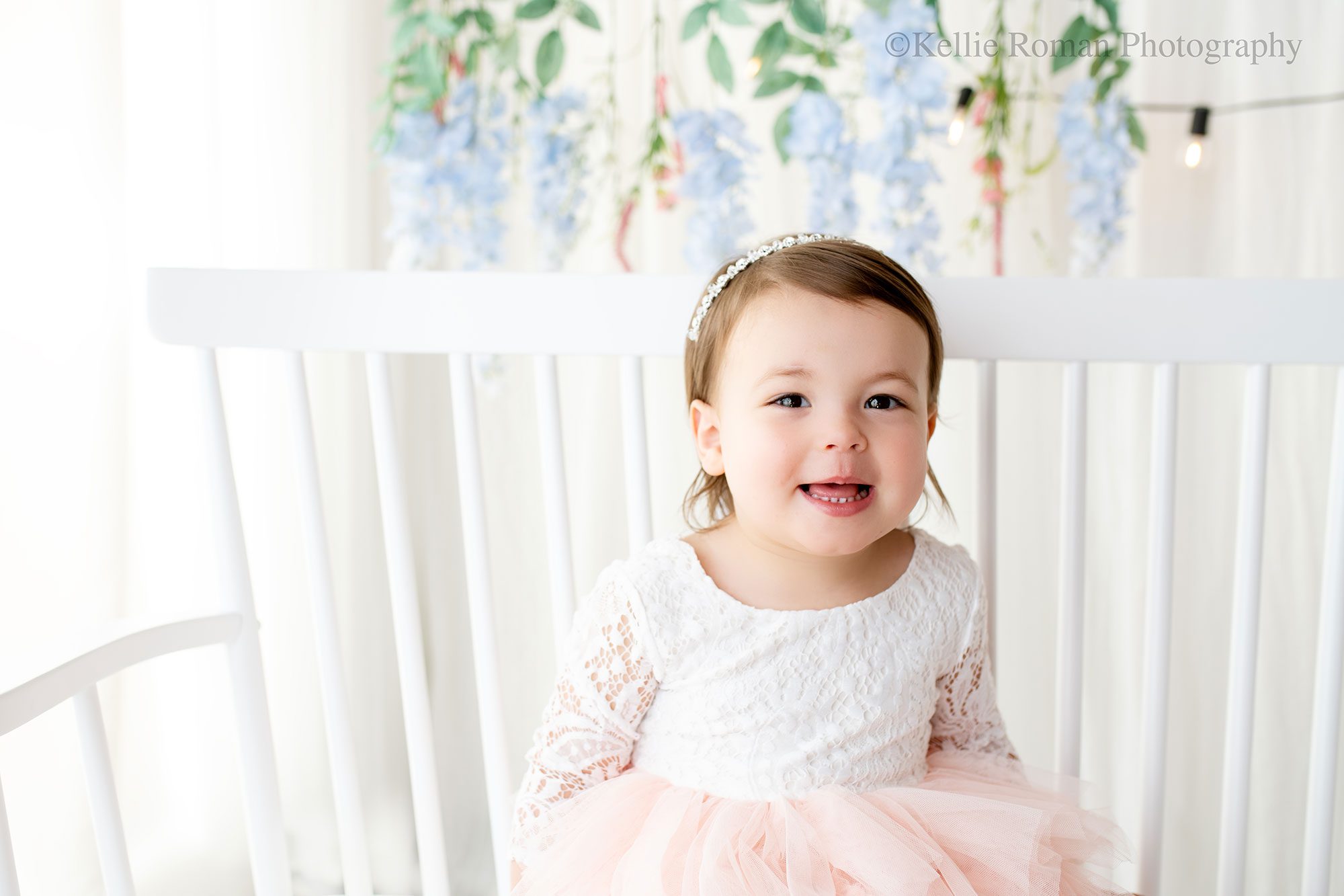 airy milestone Milwaukee. two year old girl is sitting on white wood bench . her dress is white and light pink, the backdrop is all white curtain with hanging flowers.
