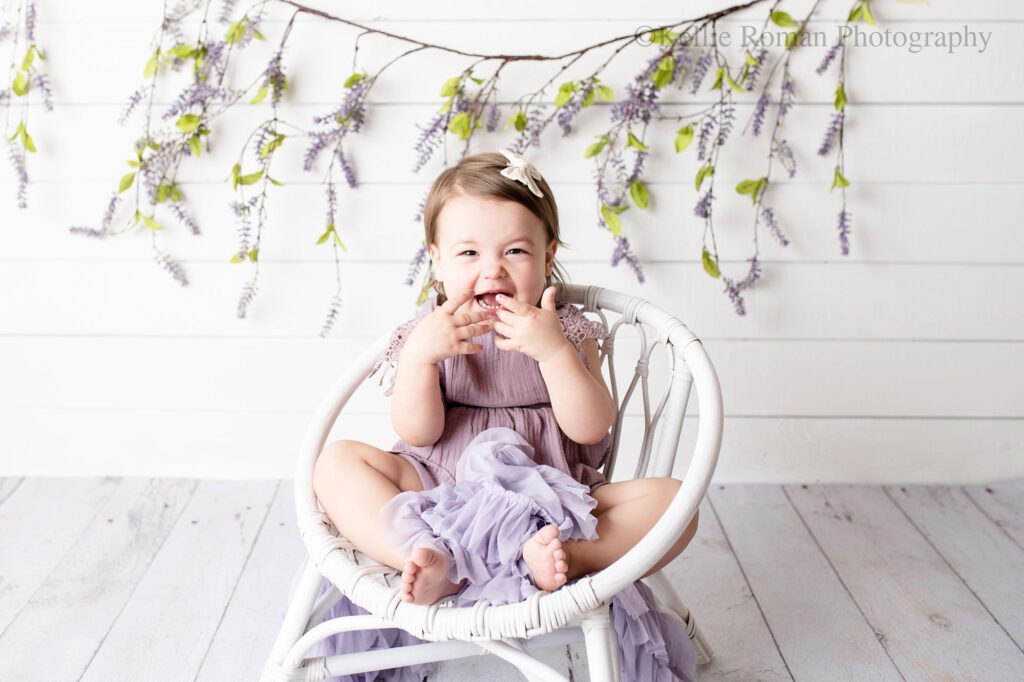 airy milestone milwaukee. two year old girl with lavender romper on sitting in white boho chair with ruffle purple fabric on her lap. backdrop is white wood with lavender flowers hanging. 