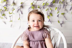 airy milestone milwaukee. two year old girl is sitting on a white wood chair. she's wearing a lavender romper with a bow clip in her hair and smiling. the backdrop is white wood with lavender flowers.