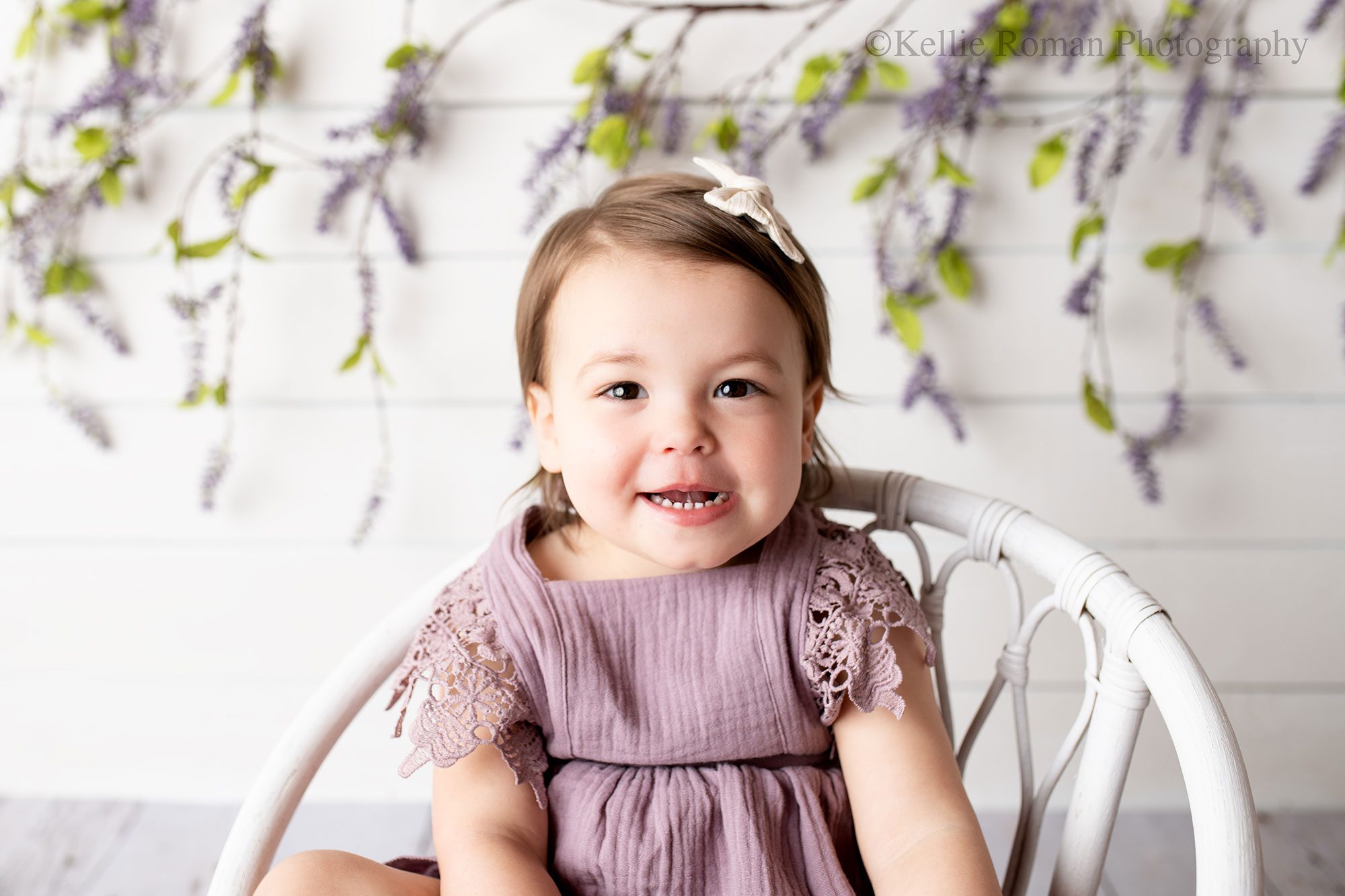 airy milestone milwaukee. two year old is sitting infront of wood backdrop with hanging lavender. she has a lavender romper on with a bow in her hair. 