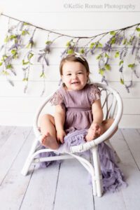 airy milestone milwaukee. two year old girl sitting on white wood chair in front of white backdrop with hanging lavender. girl has purple romper on and is smiling.