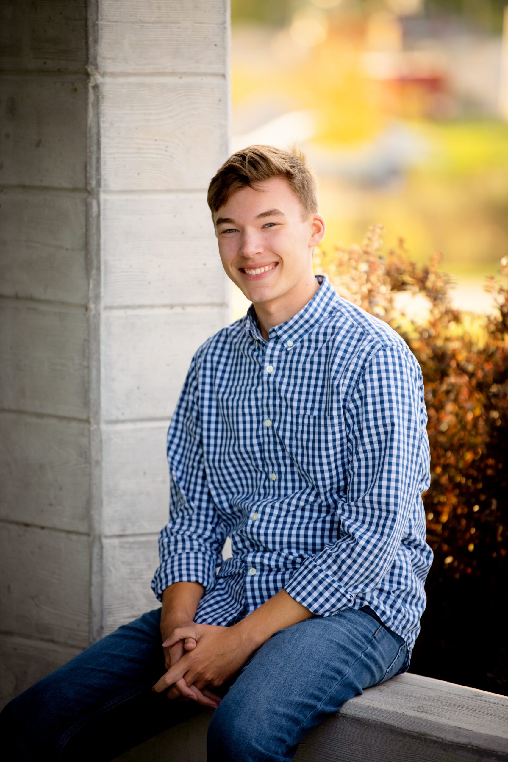 senior pictures. greendale senior boy sitting on concrete wall with blue and white checked shirt on smiling at the camera
