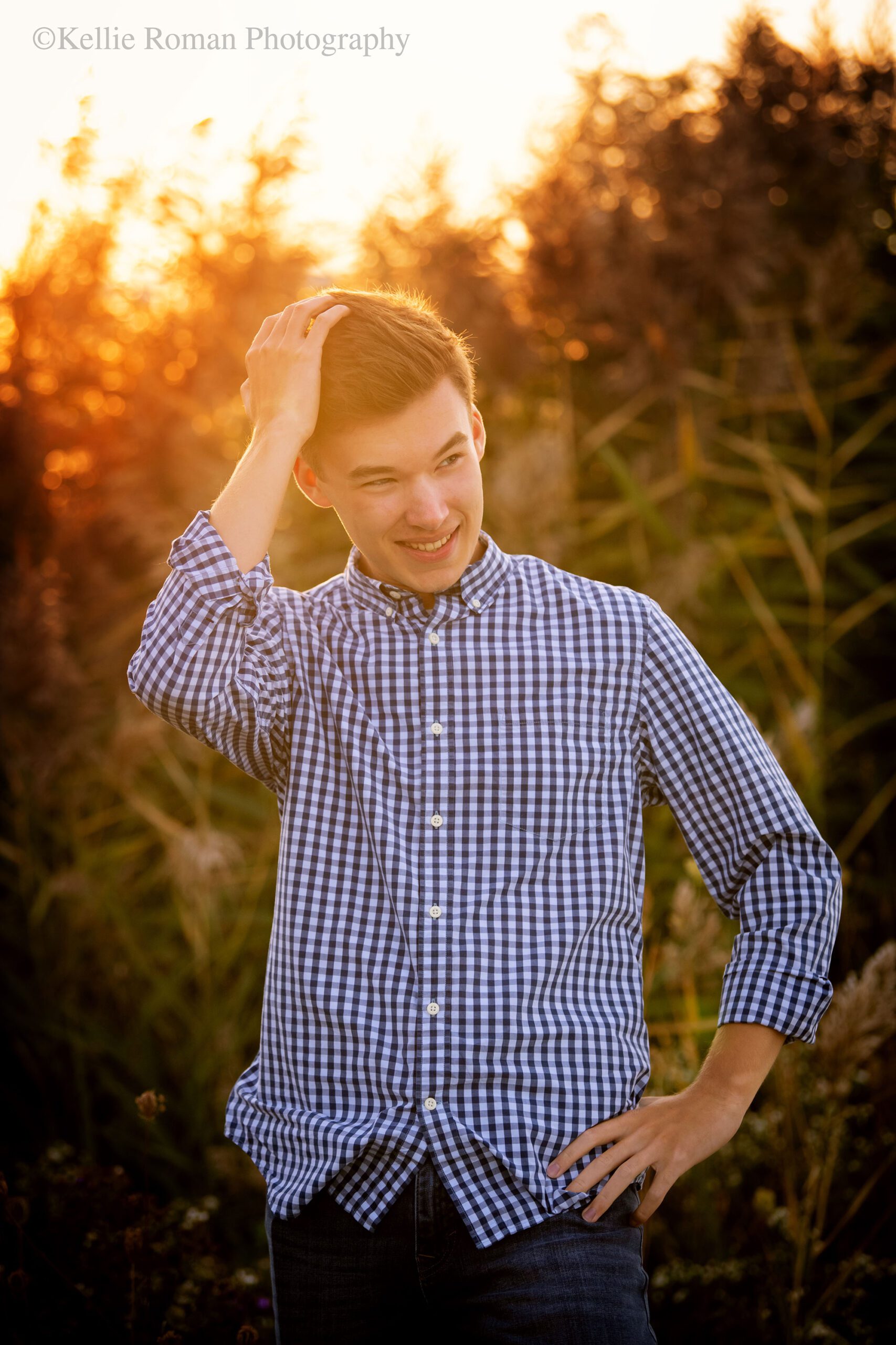 senior pictures. Greendale High School senior standing infront of tall grass with sun setting behind him. he's looking to the side and running his hands through his hair. he has a blue and white checked shirt on. 
