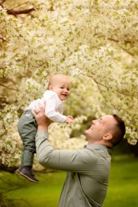 spring in milwaukee. father wearing a olive green shirt is holding his one year old son up in the air in greendale park. son is smiling and so is the father. the backdrop is green grass and trees with white flowers.
