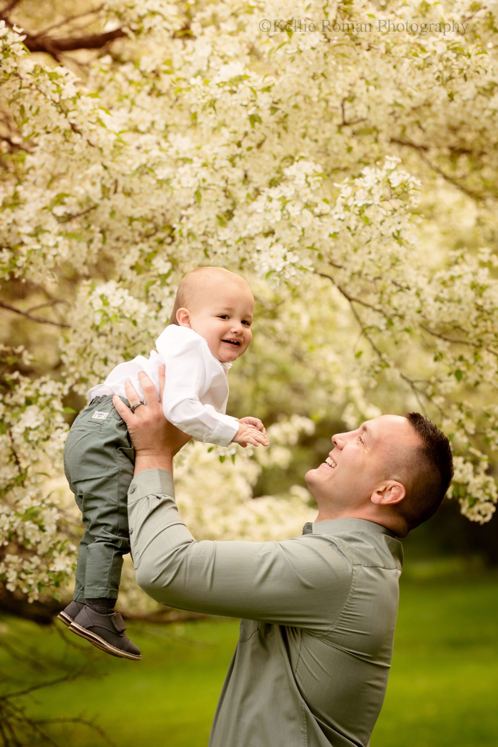 spring in milwaukee. a father is holding his one year old son up high in the air in a greendale park. there are trees with flowering trees behind them, and they are both smiling at each other. 