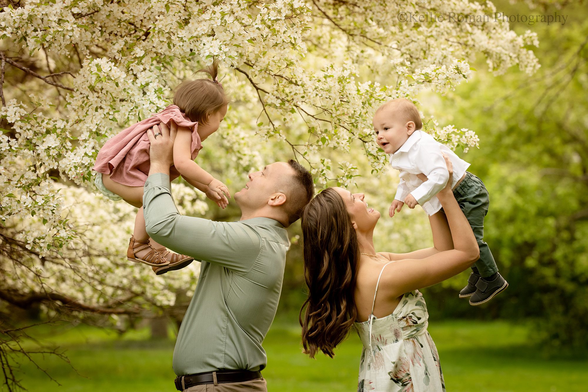 spring in milwaukee. a husband and wife are standing back to back in greendale park. mom is holding toddler son in the air, and dad is holding toddler daughter in the air. they are looking at their children and smiling. trees are full of flowers behind them
