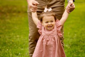 spring in milwaukee. close up shot of a dad hands holding onto his toddler daughters hands while she walks. little girl has a blush pink dress on. background is green grass and dads legs.