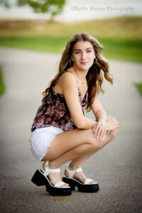 high school senior photographers. teenage girl wearing shorts and a black and purple flower tank top is crouching in roadway. she has her arms resting on her knees and is smiling while the wind blows through her hair.