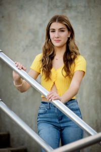 high school senior photographers. teenage girl standing on brick stairs holding onto a metal railing. she has a yellow crop top on and jeans. she has long brown hair.