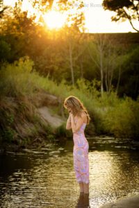 high school senior photographers. teenage girl standing in water on beach in racine. she has on a pink and purple summer swirl dress. she's looking down while running her hands through her hair. the sun is setting behind her over the hill.