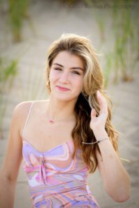 high school senior photographers. teenage girl with pink and purple swirl dress on is sitting on racine beach in sand. she has her hand running through her long hair.