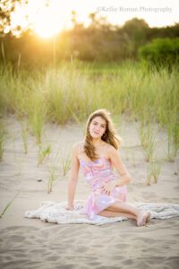high school senior photographers. teenage girl is sitting on a tan blanket on the sand on beach. girl has a pink and purple summer dress on and is barefoot. she has long hair and the sun is setting behind her.