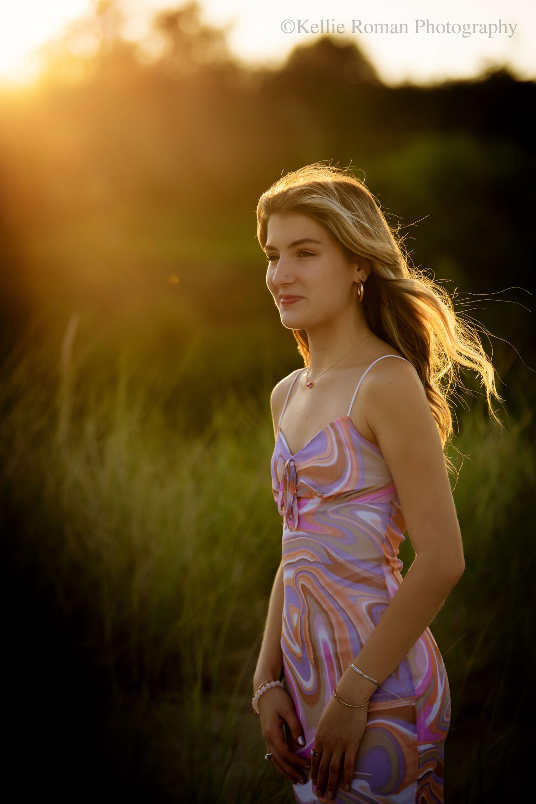 high school senior photographer.senior girl is on racine beach. she has on a pink and purple swirl tank top dress and is looking off in the distance with the golden sun setting behind her. her hair is light up in the sunlight.