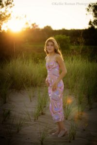high school senior photographers. teenage girl is standing on beach with grass around her. she has a pink and purple swirl summer dress on. the sun is golden as it's setting behind her and her hair is light up gold.