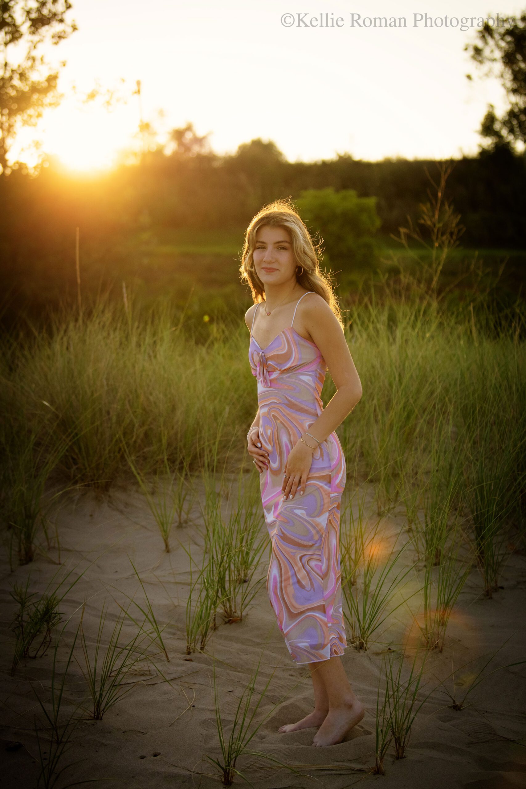 high school senior photographer. racine high school girl is standing with pink and purple dress on in the sand on the beach. the sun is setting behind her and it's glowing gold. her hair and beach grass is lite up from the sun.