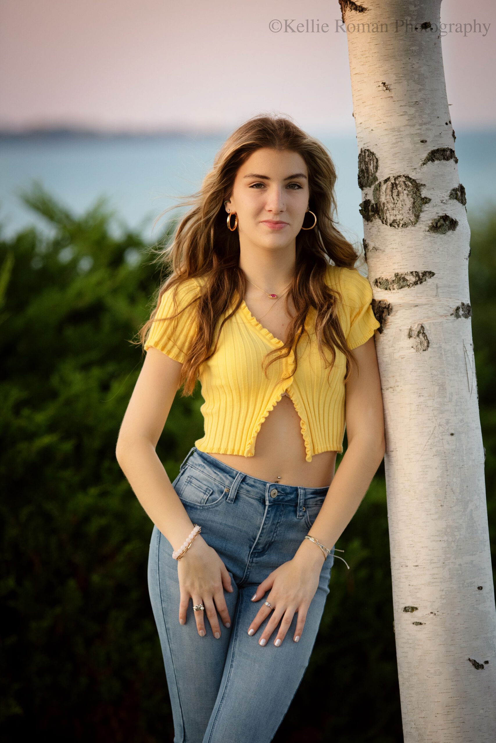 high school senior photographer. high school girl wearing a yellow crop top and jeans is leaning against a birch tree with the wind blowing through her hair. the sky is pink behind her
