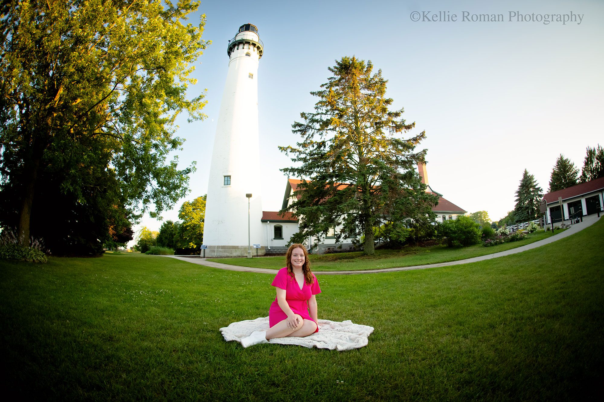 oak creek senior photography. high school senior girl wearing a hot pink dress is sitting on knit blanket in front of a white lighthouse in racine.
