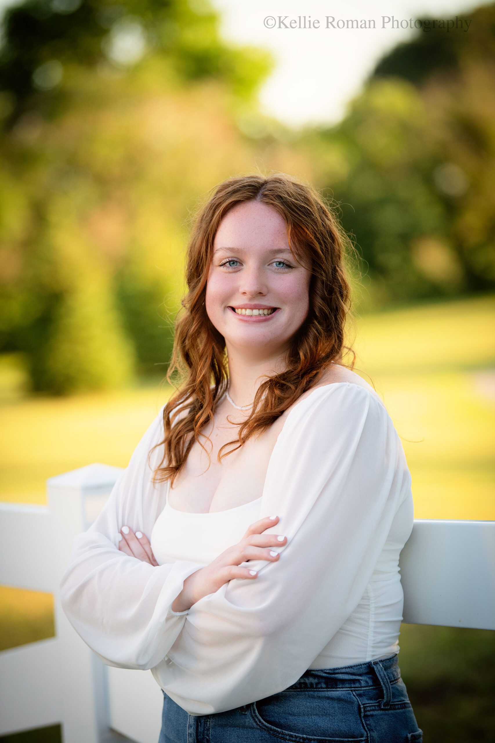 oak creek senior photography. high school senior girl is in a racine park leaning against a white fence. she has on a jeans and a white flowing top. she has her arms crossed and is smiling. she has long red curly hair. 

