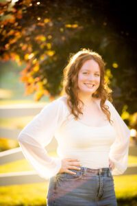 oak creek senior photography. high school girl standing with hands on hips in golden sunlight. she has jeans and white flowing top on with long red curly hair.