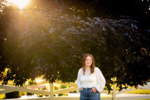oak creek senior photography. high school senior girl standing in racine park with jeans and a white flowing top on. she has her hands in pockets and is in front of a white fence and large dark tree with sun shining through the leaves.