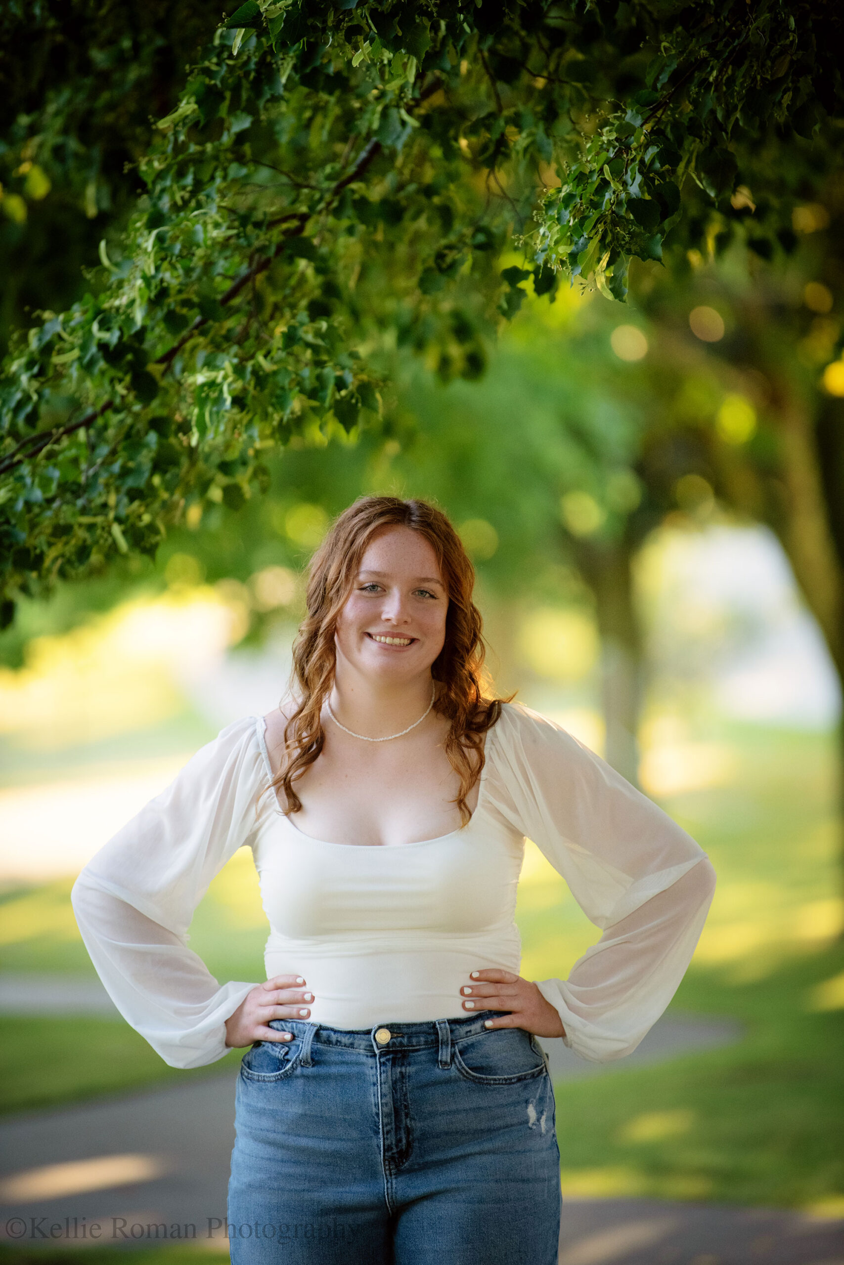 Oak Creek senior photography, high school senior girl standing in park with green trees behind her. she has jeans on and a white top. she's standing with her hands on her hips and smiling. her hair is curly, long, and red. 