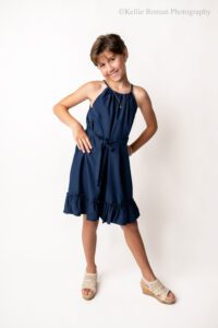 milwaukee birthday photos. ten year old girl in milwaukee photo studio. theres a white backdrop, and the girl has a navy dress on with tan sandals. she's standing with her hands on her hips and smiling.