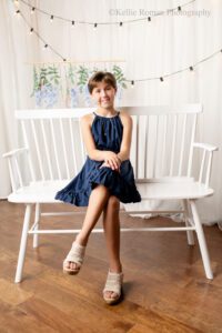 milwaukee birthday photos. ten year old girl sitting on a white bench in milwaukee photo studio. she has a navy dress on with tan sandals and her legs are crossed. the background is white curtains with lights and purple flowers hanging.