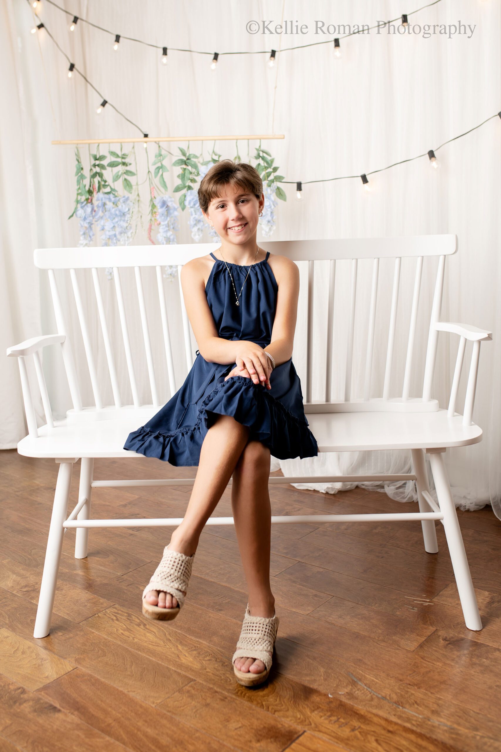 milwaukee birthday photos. 10 year old girl sitting on a white bench with legs crosses. she has a navy dress with tan sandals on. the backdrop is white curtains with lights, and purple hanging flowers.