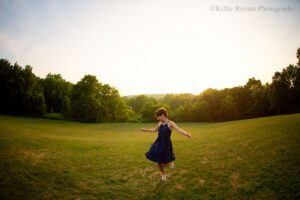 milwaukee birthday photos. ten year old girl is onto of a hill of green grass spinning around. there are trees in the distance. she's wearing a navy dress with tan sandals.