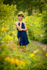 milwaukee birthday photos. ten year old girl is in milwaukee parking surrounded by tall green grass and wildflowers. she has a navy dress on and has her hand near her face while smiling at the camera.