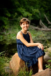 milwaukee birthday photos. young girl is sitting on large rock near pond. there are trees behind her and tall grass around her. she has a navy dress on and is crossing her legs while smiling at the camera.