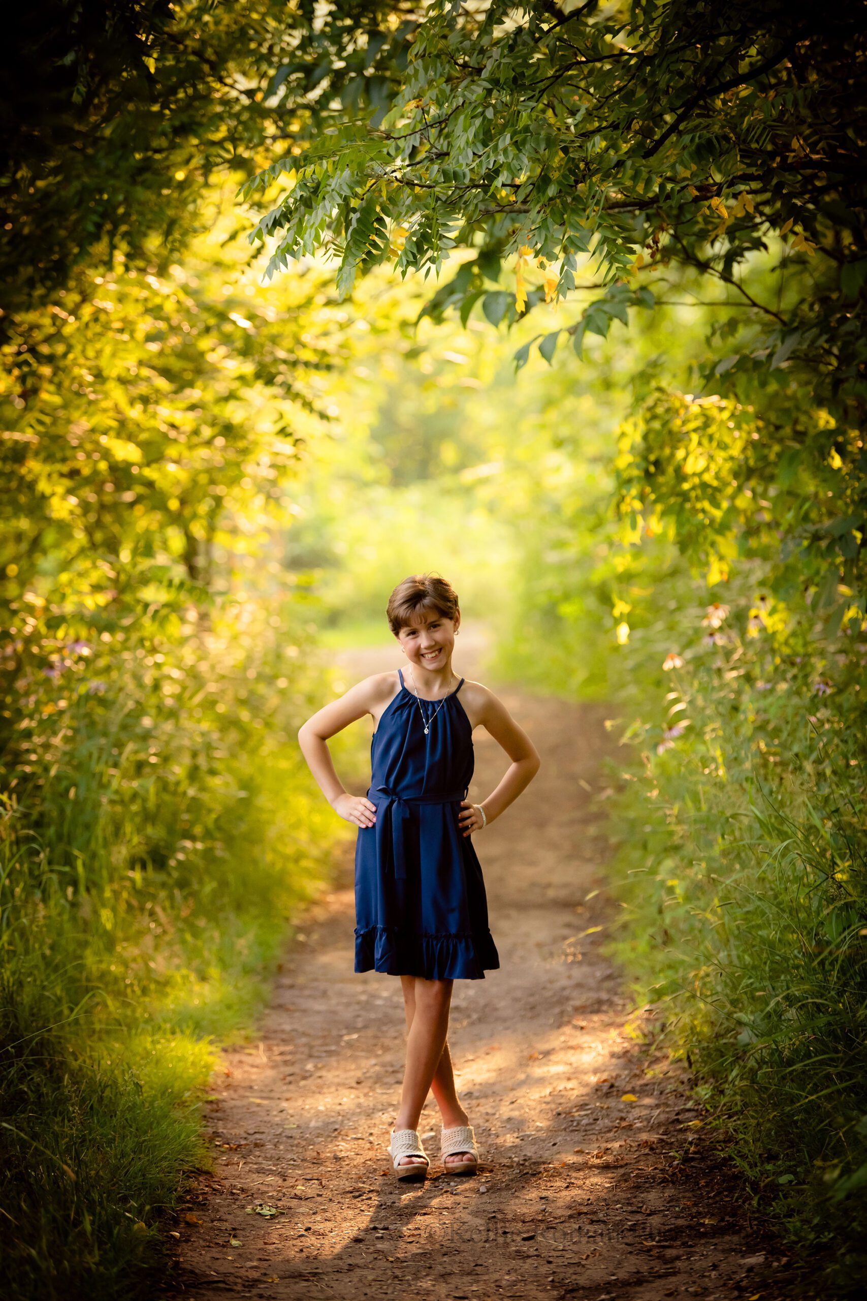milwaukee birthday photos. 10 year old girl surrounded by trees in park is standing on dirt path. she has her hands on her hips and is wearing a navy dress with tan sandals.